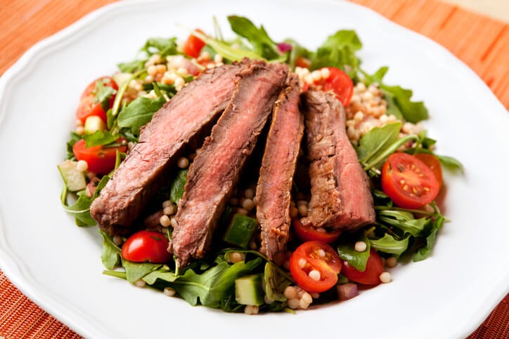 Slices of cooked steak on top of a couscous and greens salad on a plate.