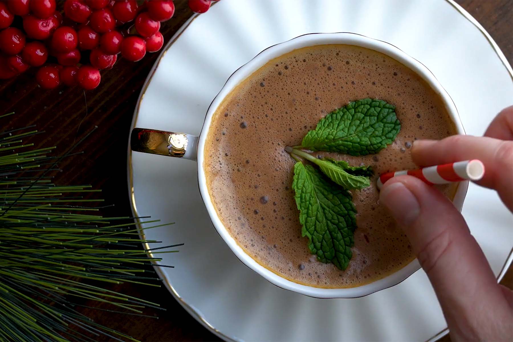 A hand adds a sprig of mint to a cup of chocolate drink, surrounded by decorative red berries and pine leaves on a wooden surface.