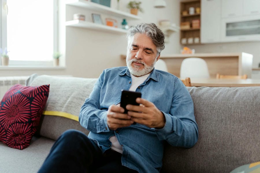 Man sitting on a couch in a living room holding and looking at a cell phone. 