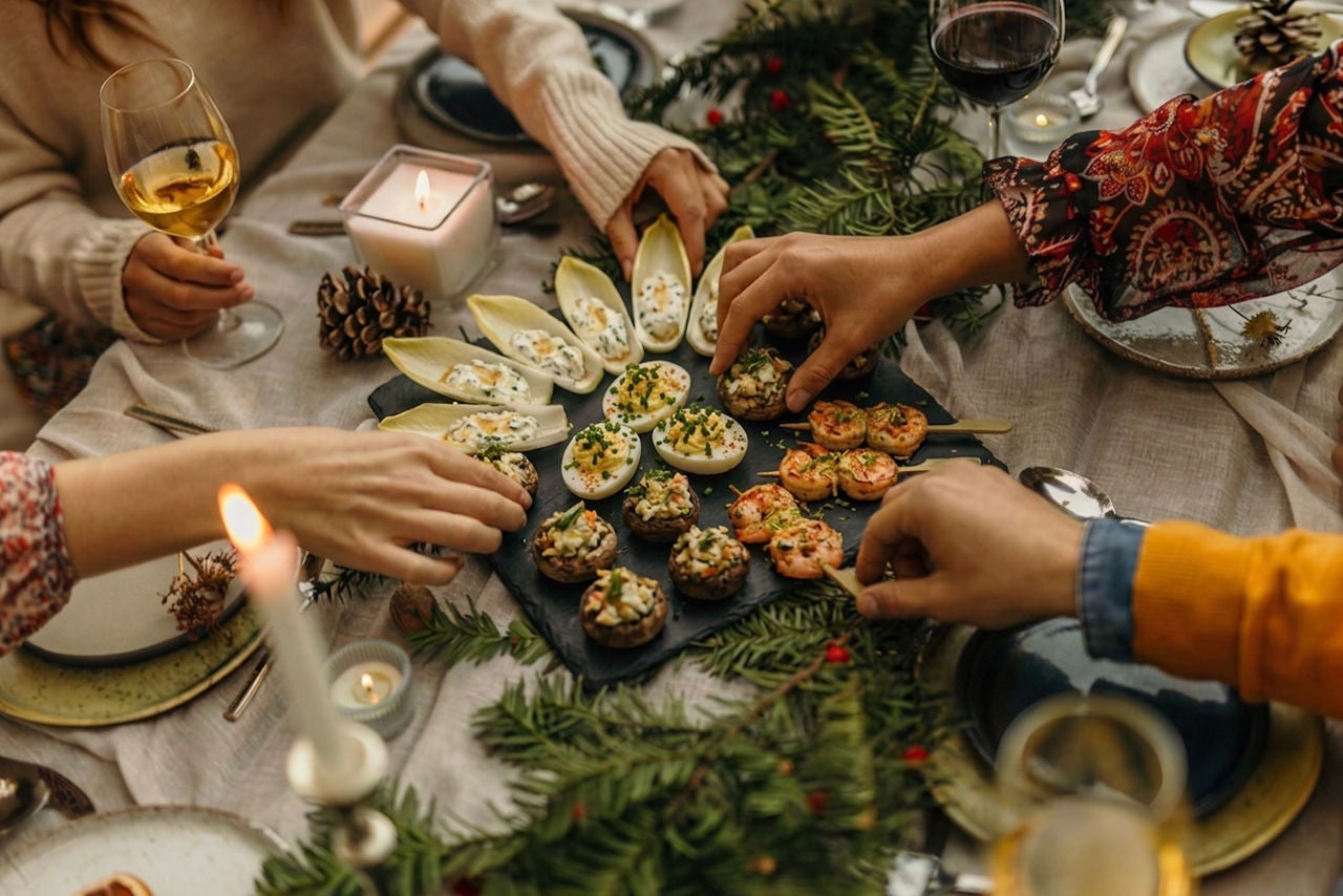 Overhead view of a set holiday table with a tray of mixed appetizers with hands reaching in to grab them. 