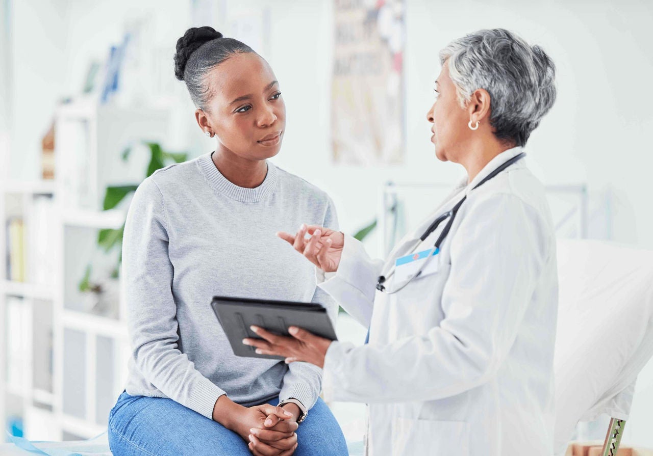 Woman sitting in brightly lit office listening intently to a doctor holding a tablet. 
