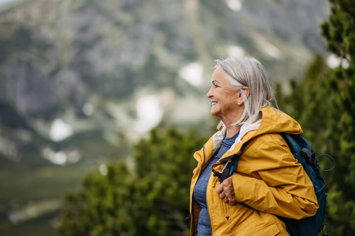 Older woman smiling while outdoors wearing hiking clothing with a scenic mountain view in the background.