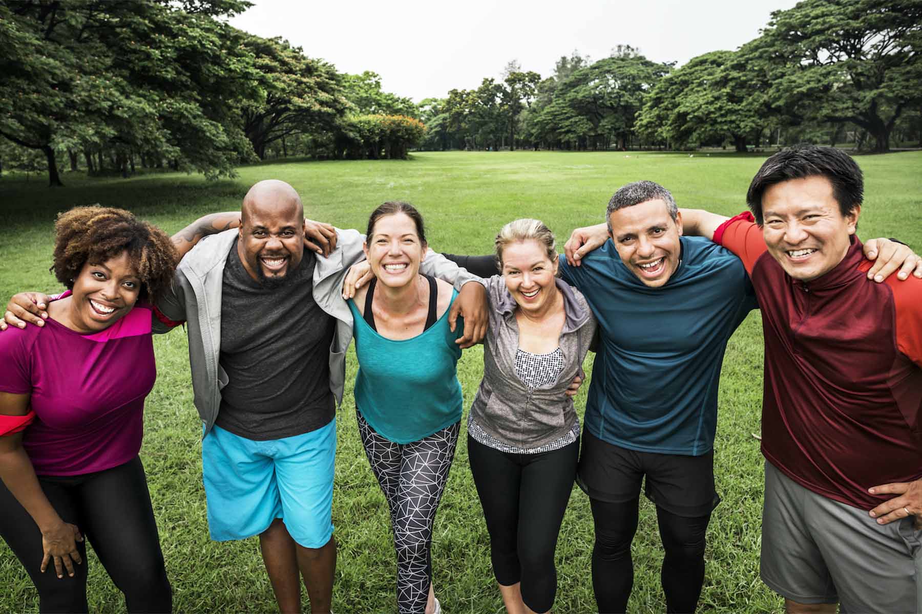 Group of six diverse friends standing together outdoors in a park, with arms around each other, smiling and laughing.