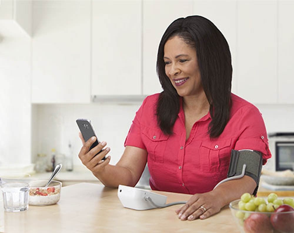 Una mujer con camiseta roja revisa su smartphone mientras usa un monitor de presión arterial en la mesa de la cocina, con frutas al fondo
