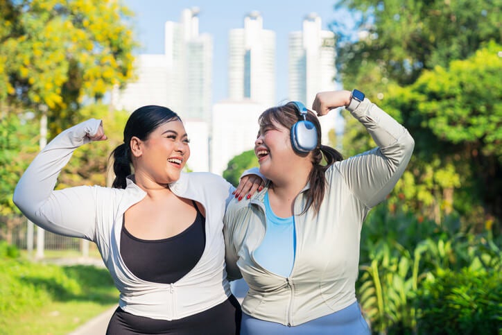 Two women wearing athletic clothing walking on an outdoor path with their arms around each other smiling and flexing their biceps.