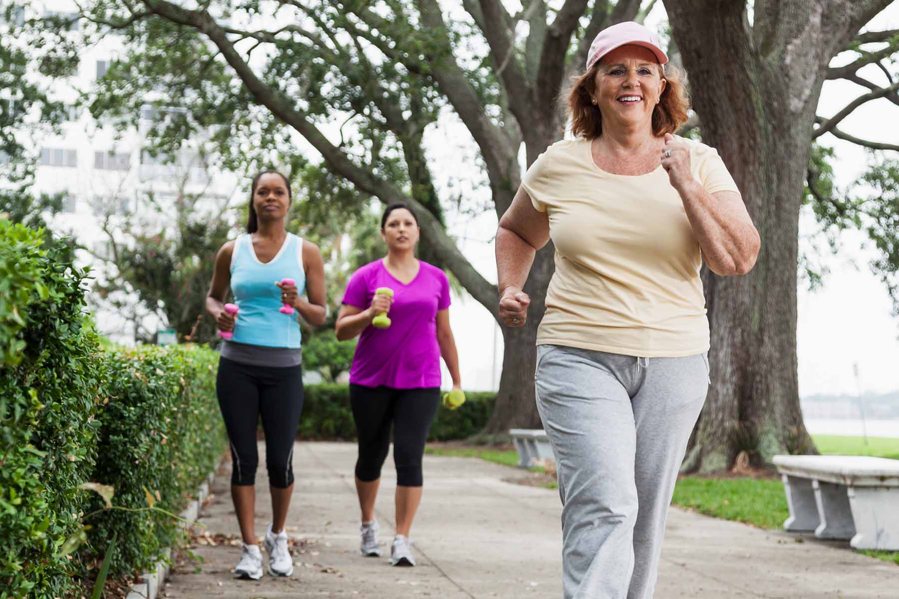 Group of women walking briskly along a scenic tree-lined path in a park, focusing on fitness and enjoying the outdoors, with lush greenery and park benches in the background.