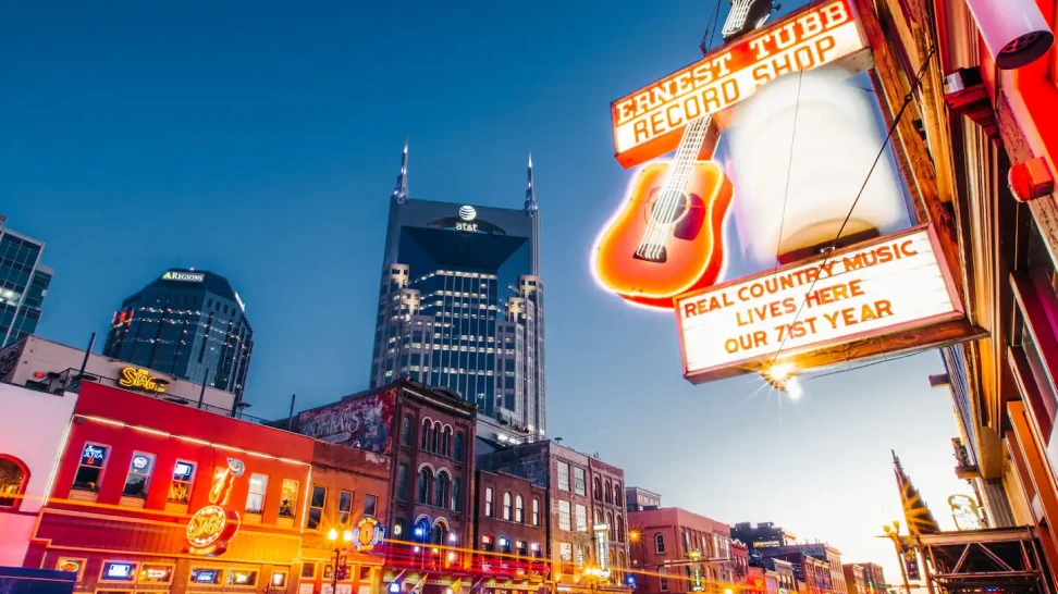Neon-lit street at dusk with a large guitar sign above a music shop and modern skyscrapers in the background