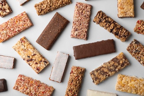 Various granola and energy bars with different textures and toppings scattered on a light gray surface.