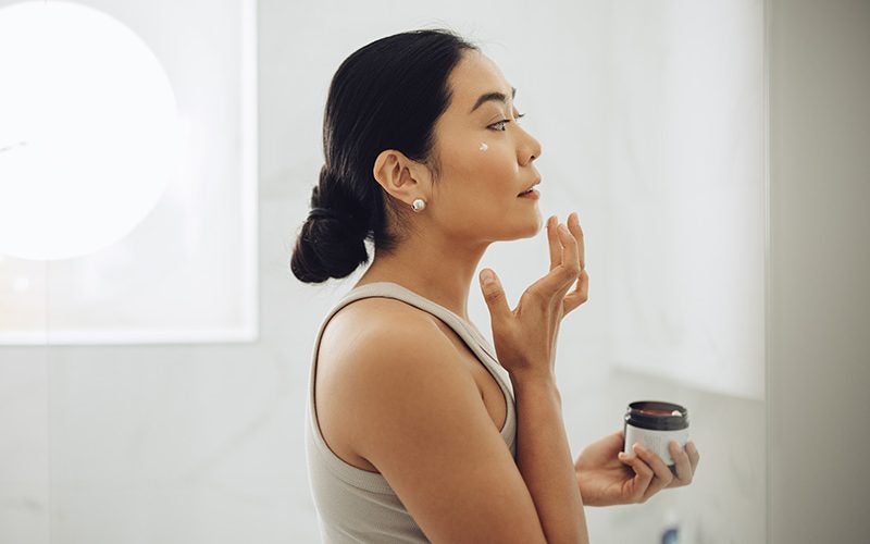 Woman applying facial moisturizer while looking in the mirror.