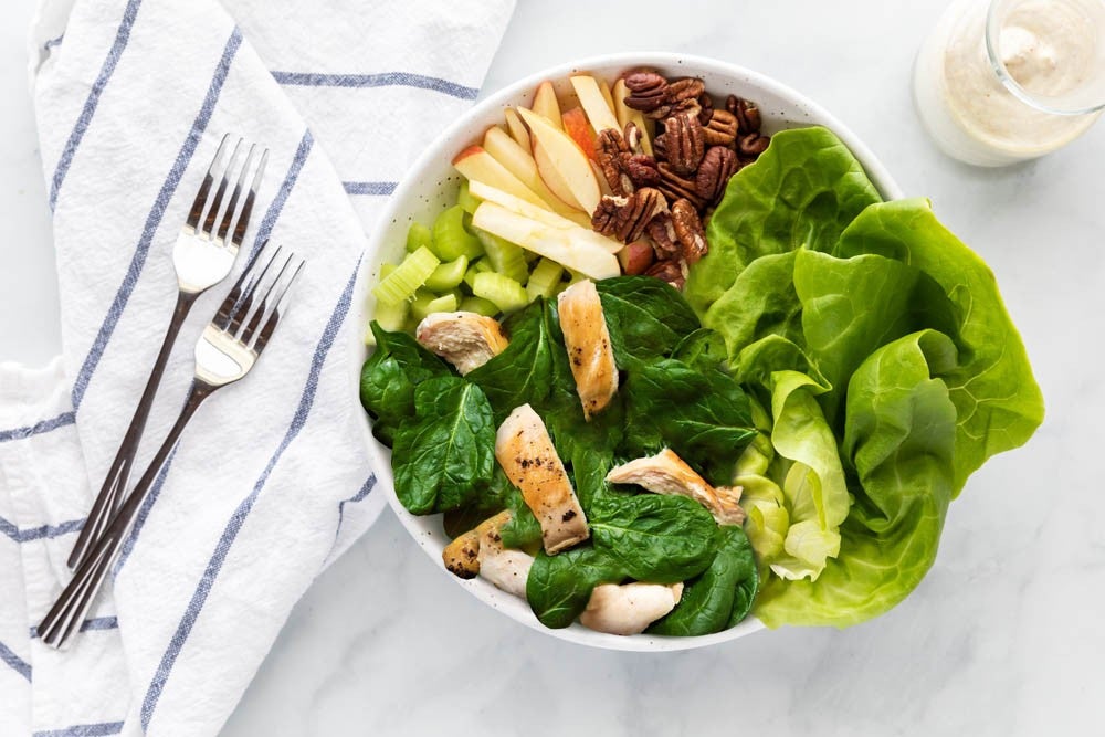 An overhead view of a salad bowl filled with greens, grilled chicken pieces, sliced apples, and pecans, accompanied by a white cloth and utensils on a marble surface.