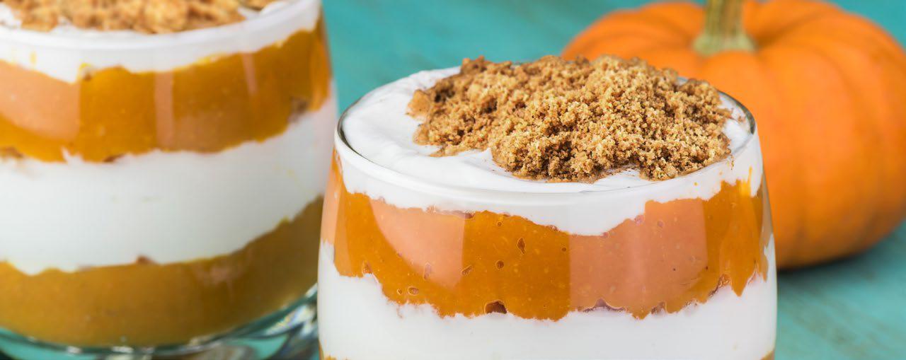 Pumpkin yogurt parfait in a glass with a pumpkin in the background on a light blue surface.