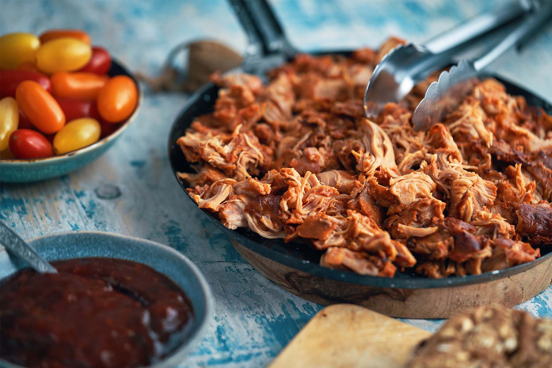 Skillet of shredded barbecue chicken with tongs, surrounded by grape tomatoes, barbecue sauce, and bread