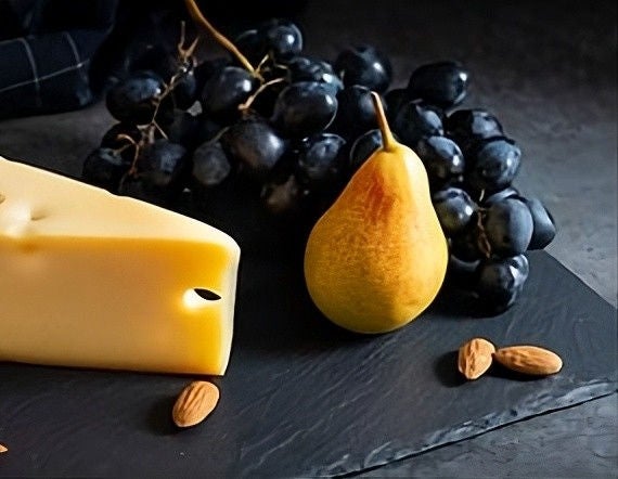 A block of cheese next to a pear and a bunch of grapes with a few almonds on a black table.