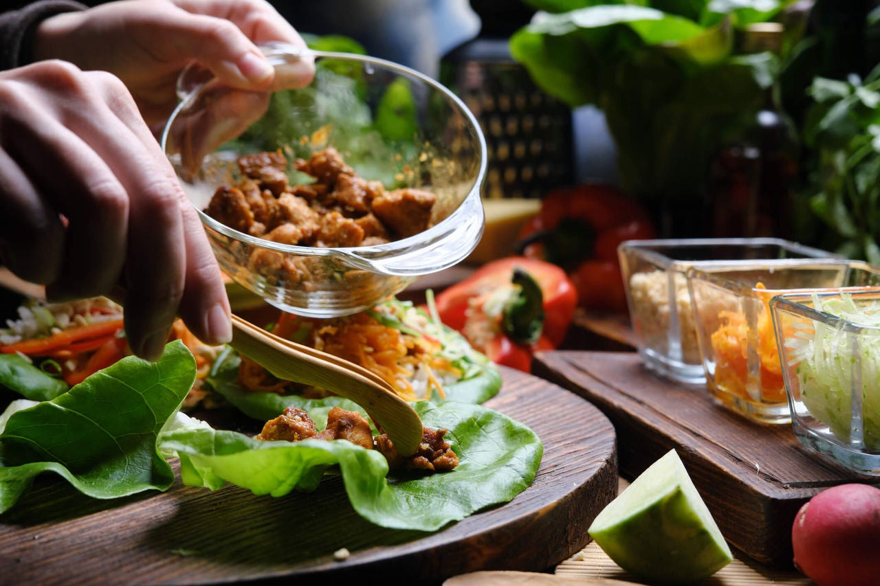 Hands placing cooked chicken onto a lettuce leaves with prepared toppings in the background.