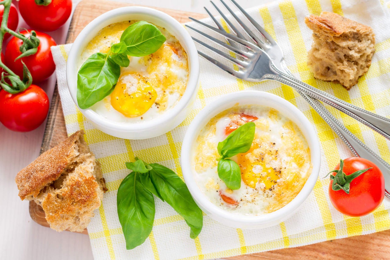 Ramekins with baked egg topped with basil, tomatoes and bread next to them. 