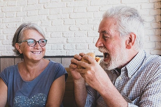 Hombre mayor comiendo un sándwich junto a una mujer con gafas sonriendo, frente a una pared de ladrillo blanco