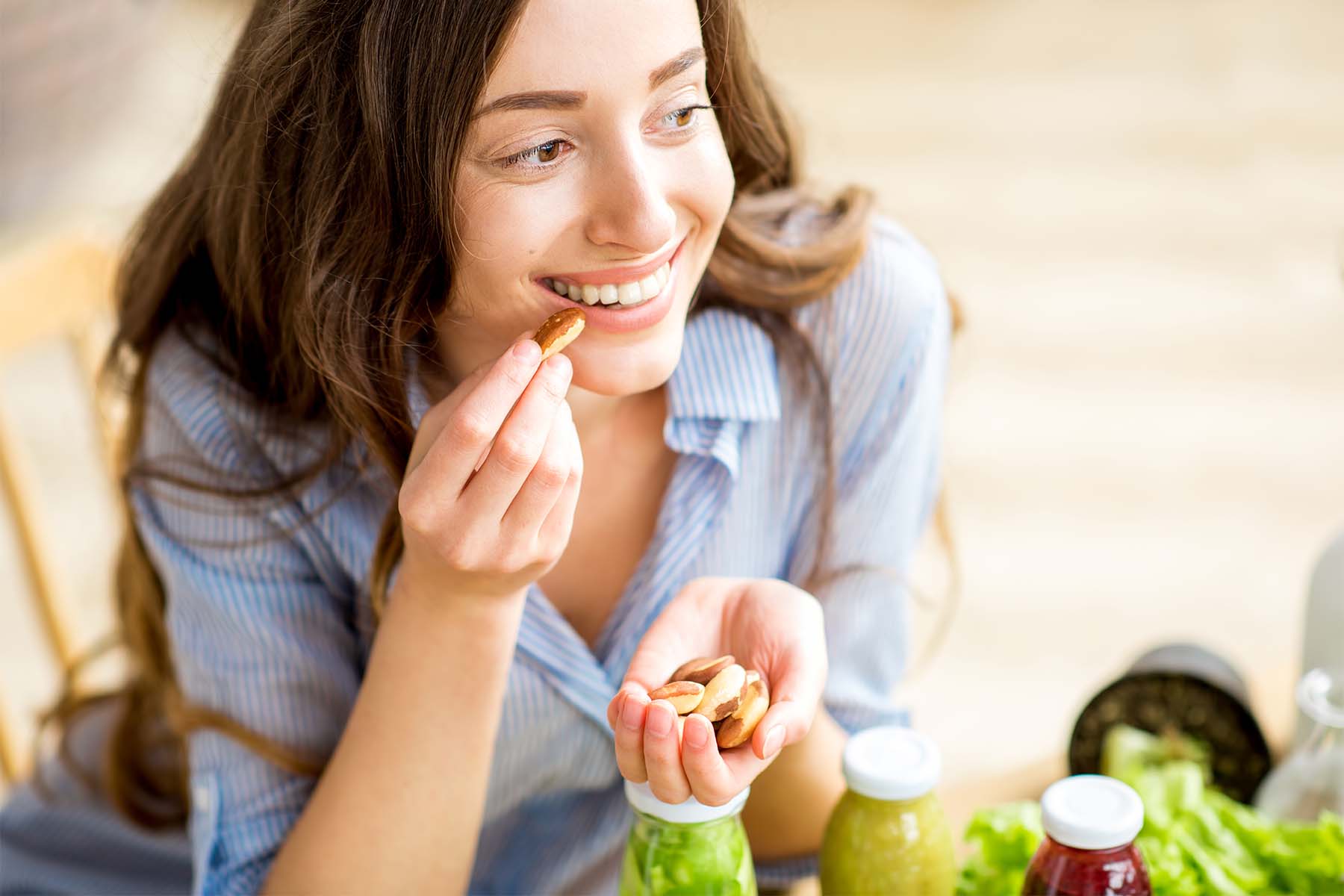 A woman smiling while holding a handful of nuts, sitting at a table with healthy snacks and drinks in the background.