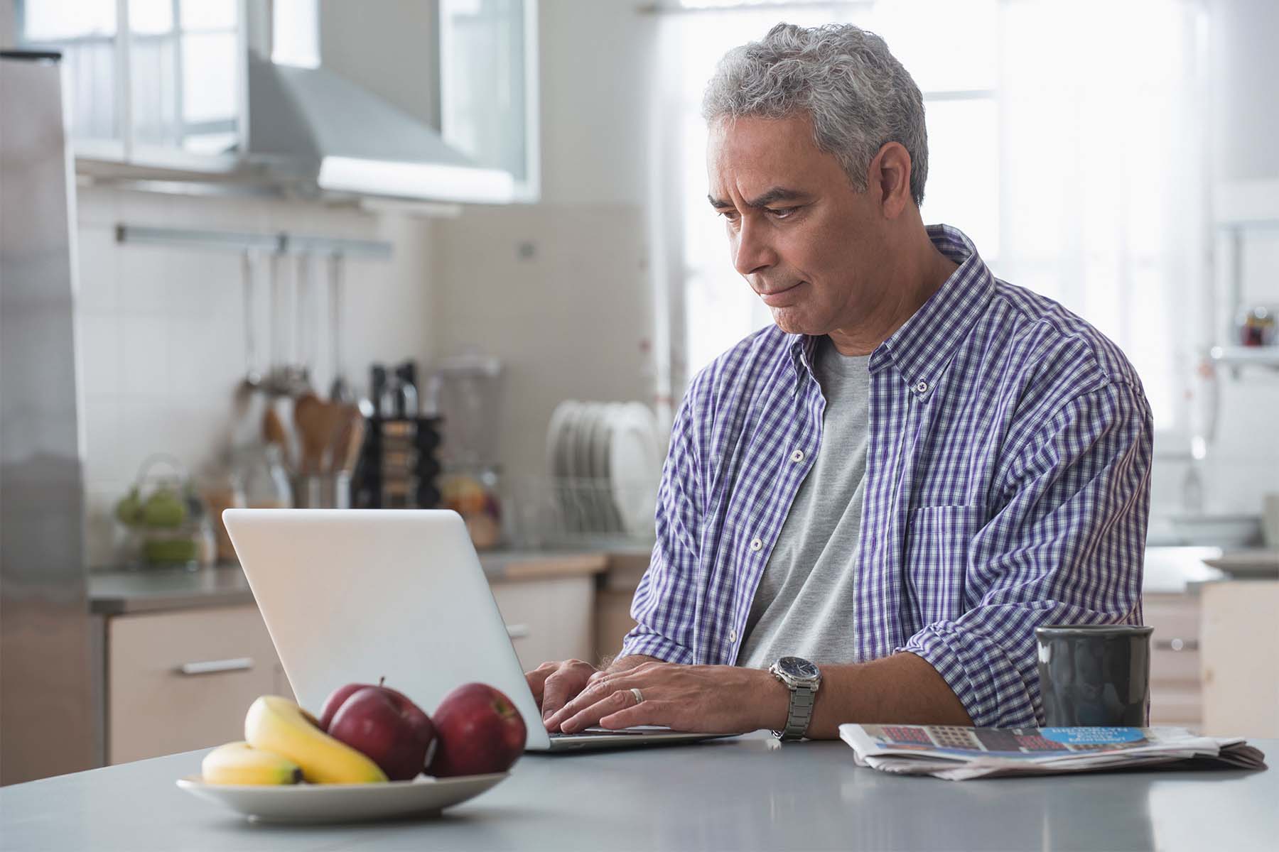 Focused older man using a laptop in a kitchen, with coffee and fruit nearby.
