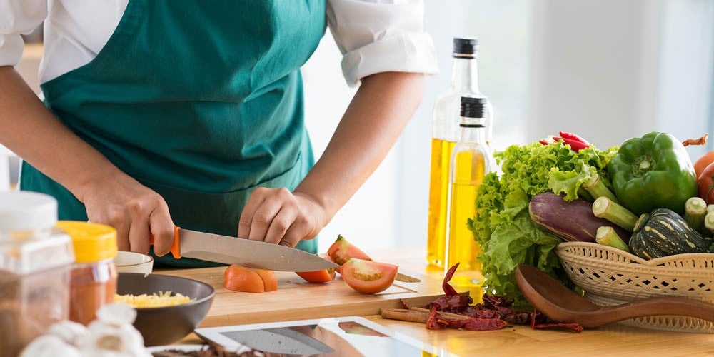 Persona con un delantal verde cortando tomates sobre una tabla de madera, rodeada de varios vegetales e ingredientes de cocina, con botellas de aceite al fondo