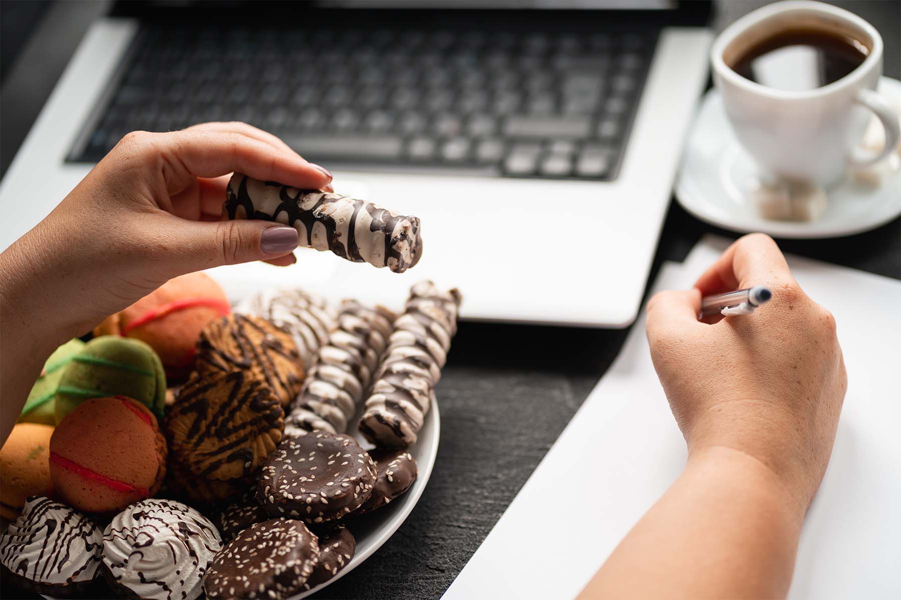 Person holding a chocolate-covered pastry while writing on paper, with a plate of assorted sweets, a laptop keyboard, and a cup of coffee on the desk.