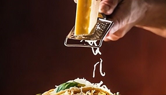 Hand holding shredder and shredding a block of cheese with cheese falling onto a plate of food.