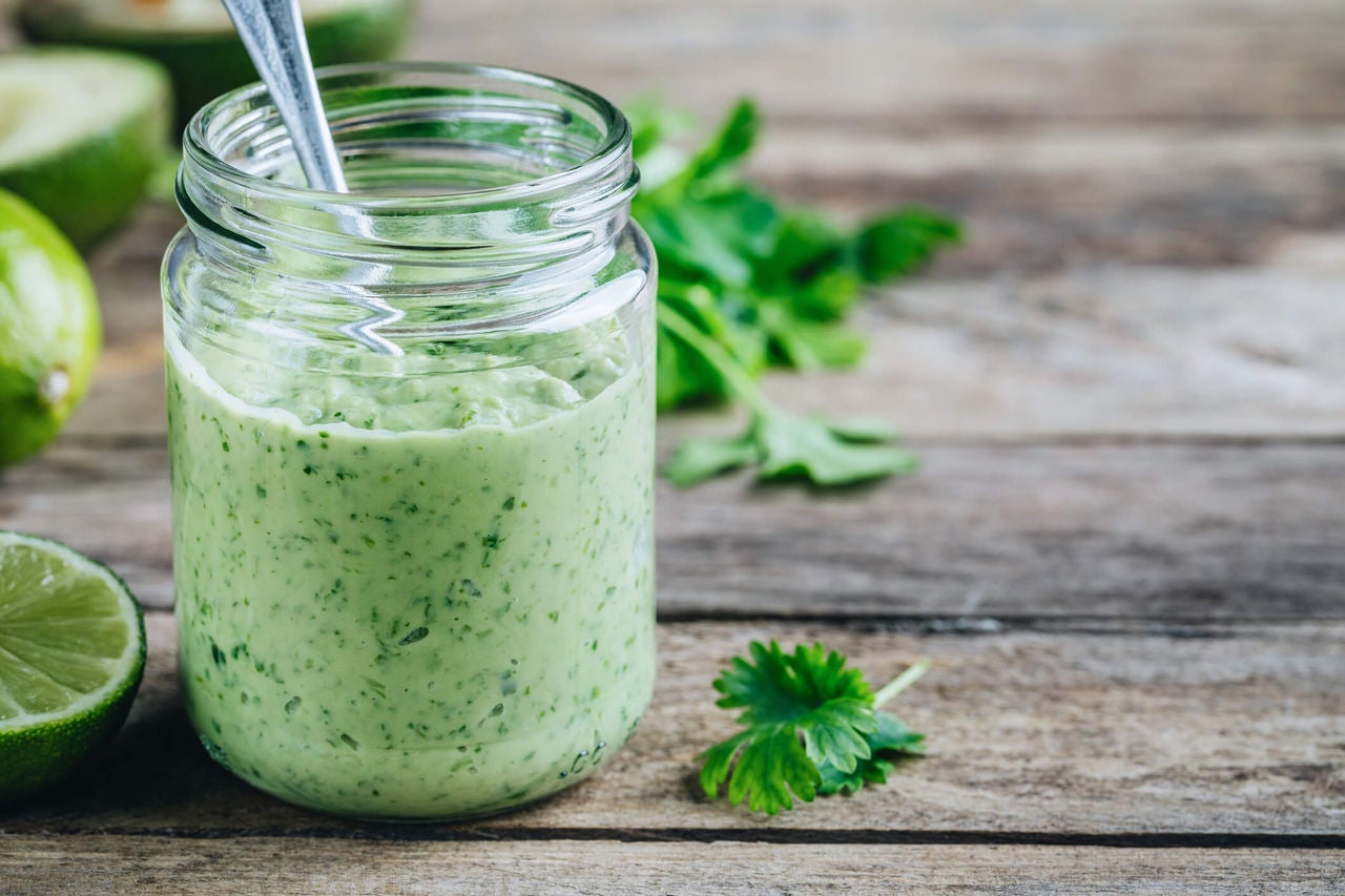 Creamy green avocado dressing with herbs in a small jar next to cilantro and lime wedges.