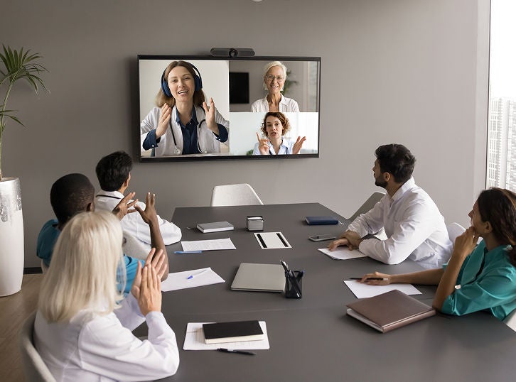 Group of healthcare professionals seated in a conference room participating in a video conference displayed on a large screen.