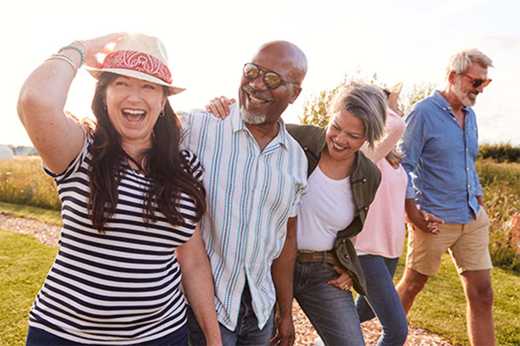 Group of four friends walking and laughing outdoors on a sunny day, surrounded by tall grass and open fields.