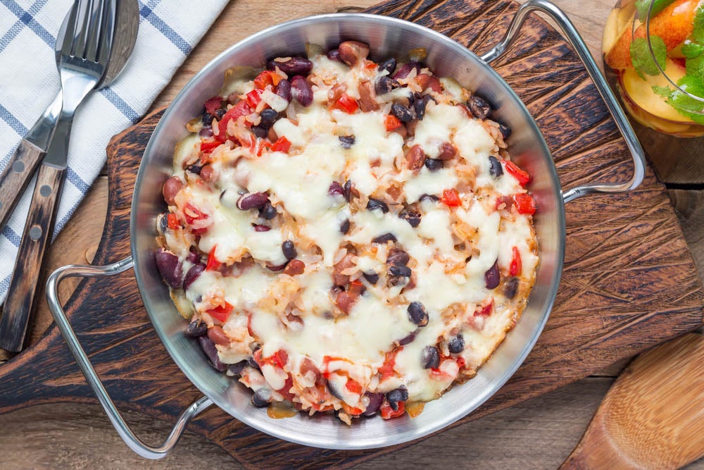 Vista superior de un platillo horneado en un molde redondo de metal con una colorida mezcla de frijoles, tomates en cubos y queso derretido, colocado sobre una tabla de cortar de madera con utensilios y un vaso al fondo