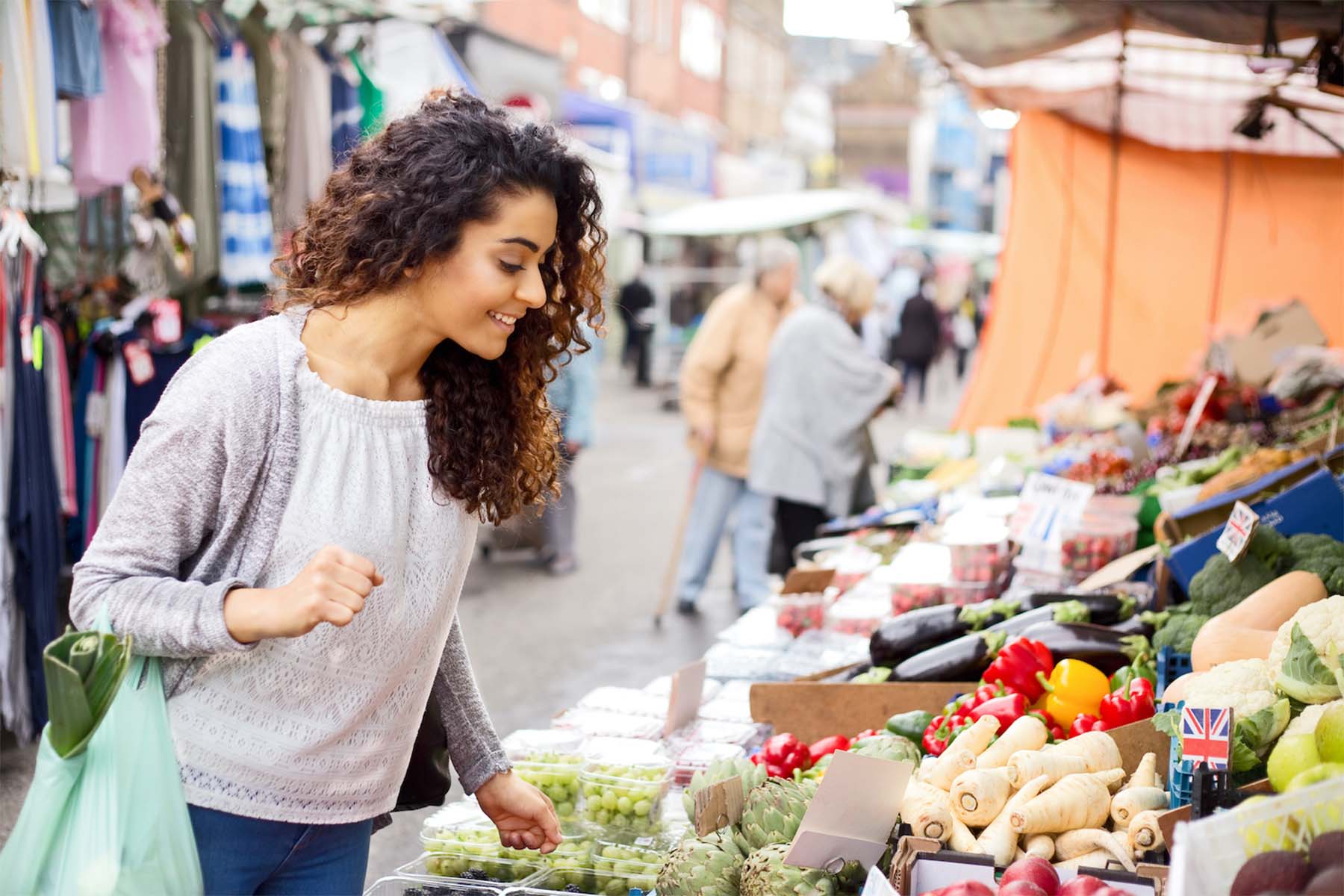 Woman shopping for fresh vegetables and fruits at an outdoor market, holding a reusable bag.