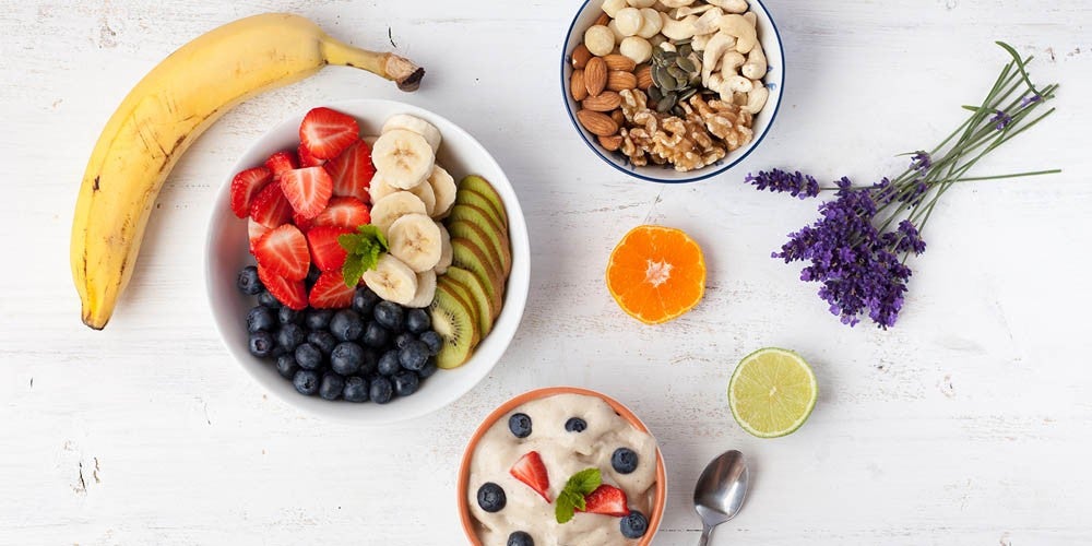 Vista cenital de una mesa con un bol de frutas mixtas incluyendo frutillas, arándanos, kiwi y banana, junto a un bol más pequeño con frutos secos y algunas ramitas de lavanda, sobre una superficie blanca