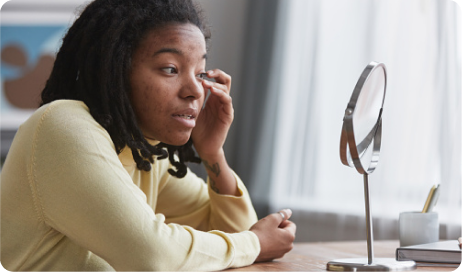  A young woman sits at a table, looking at her reflection in a mirror.