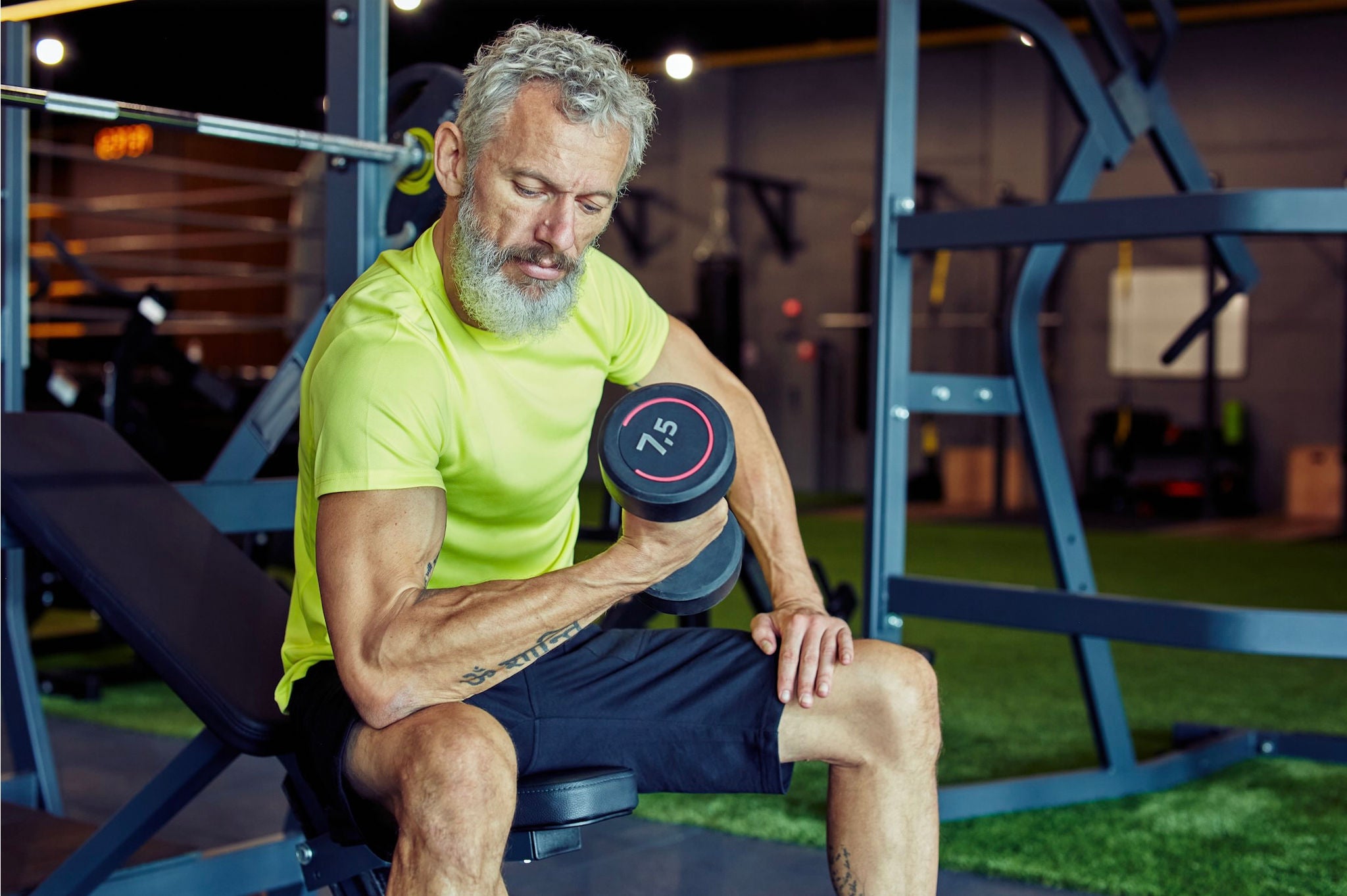 Older man wearing athletic clothing curling a dumbbell while sitting on a gym apparatus. 