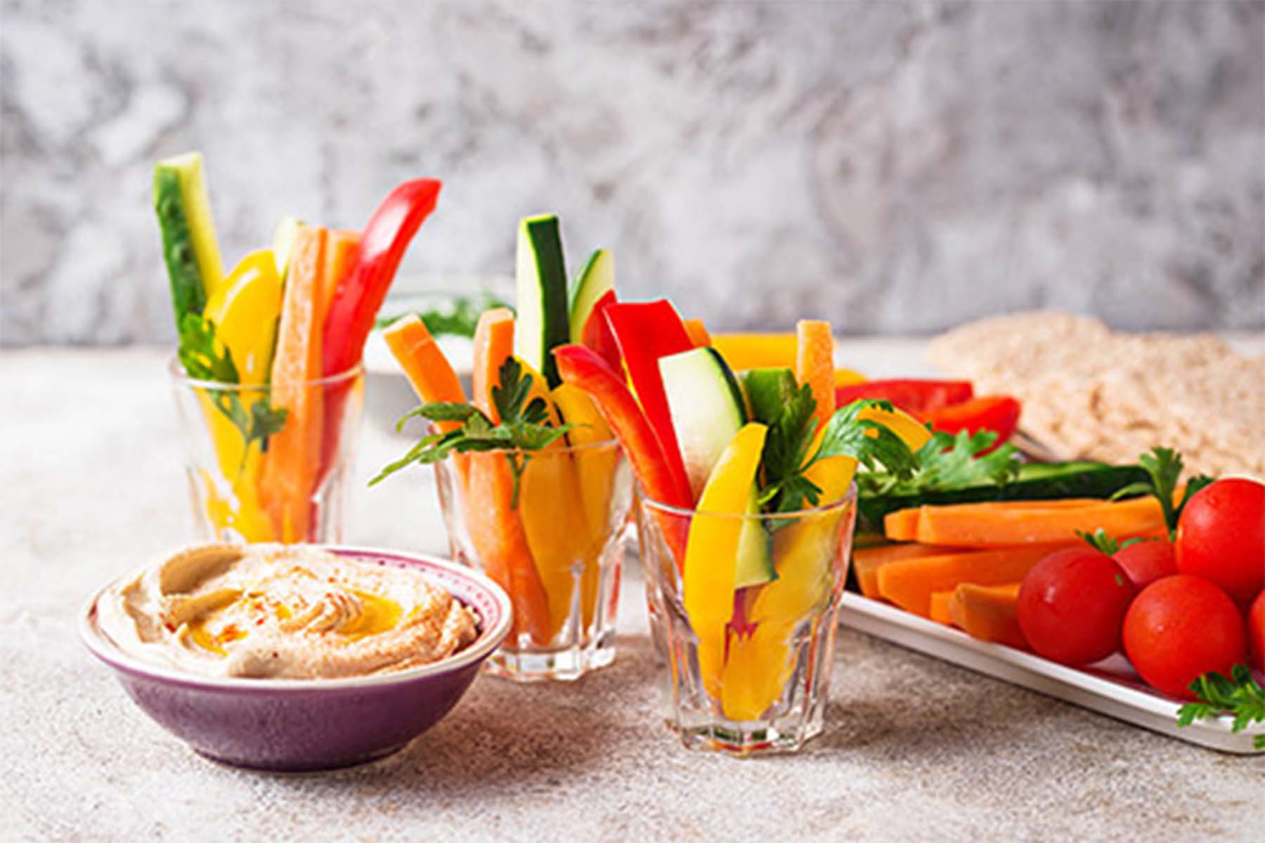 Colorful vegetable sticks in glasses, including bell peppers and cucumbers, served alongside a bowl of hummus and a platter of cherry tomatoes and additional veggies.