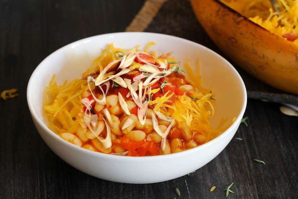 Bol de pasta cubierto con queso rallado, tomates en cubos y repollo, con una calabaza asada al fondo sobre una superficie oscura