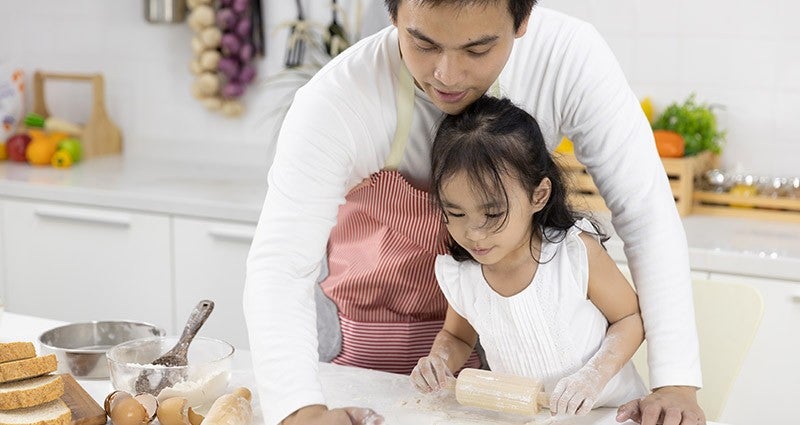 A parent and child are engaged in baking in a kitchen, with the parent guiding the child as they roll out dough on a countertop, surrounded by baking ingredients and utensils.