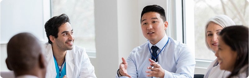 A group of five people engage in conversation in a boardroom, wearing a mix of business casual and medical attire.