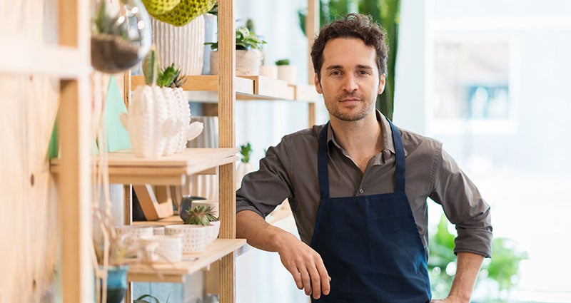 A young man in an apron is standing in a bright, modern shop filled with plants and home goods, smiling at the camera.