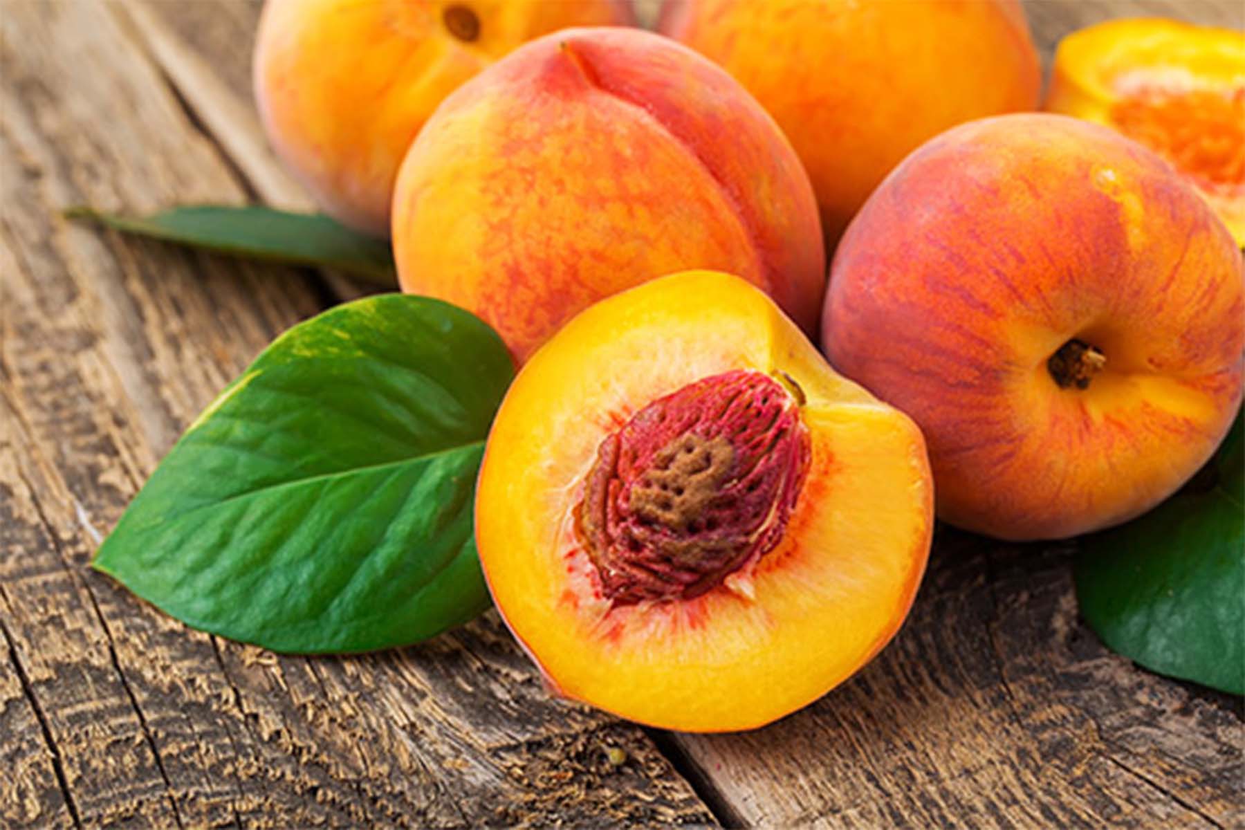 Fresh peaches, some whole and some halved, with vibrant orange and yellow skin, arranged on a rustic wooden surface with green leaves.