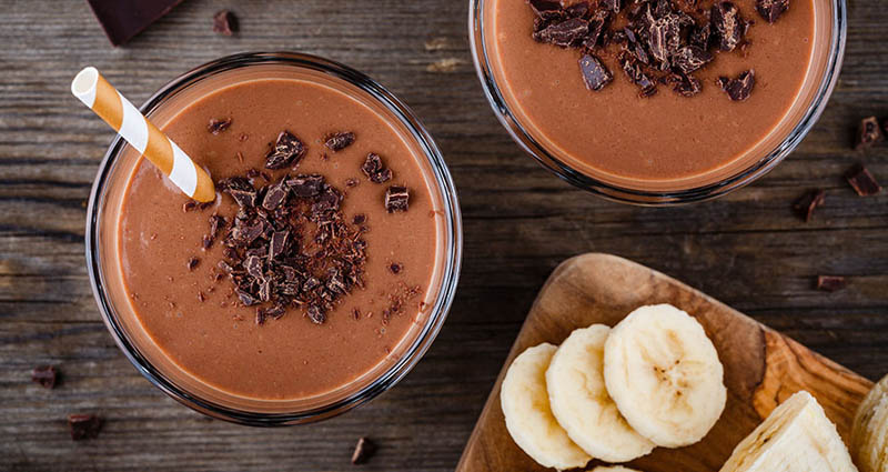 2 smoothies set on a table topped with chocolate and served with bananas on the side
