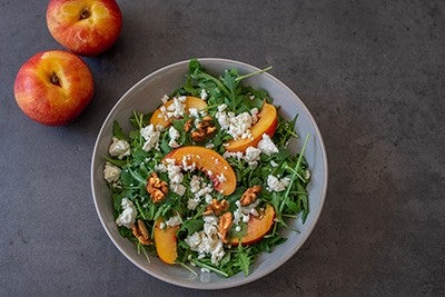 Salad with arugula, sliced peaches, walnuts, and crumbled cheese in a bowl; two whole peaches beside it on a gray surface.