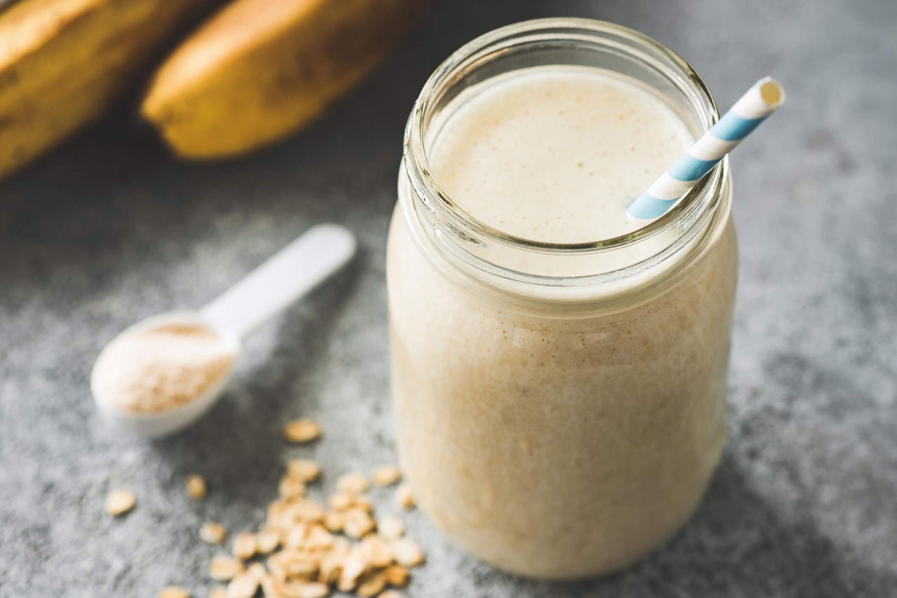 Brown smoothie with measuring spoon with powder and bananas in the background.