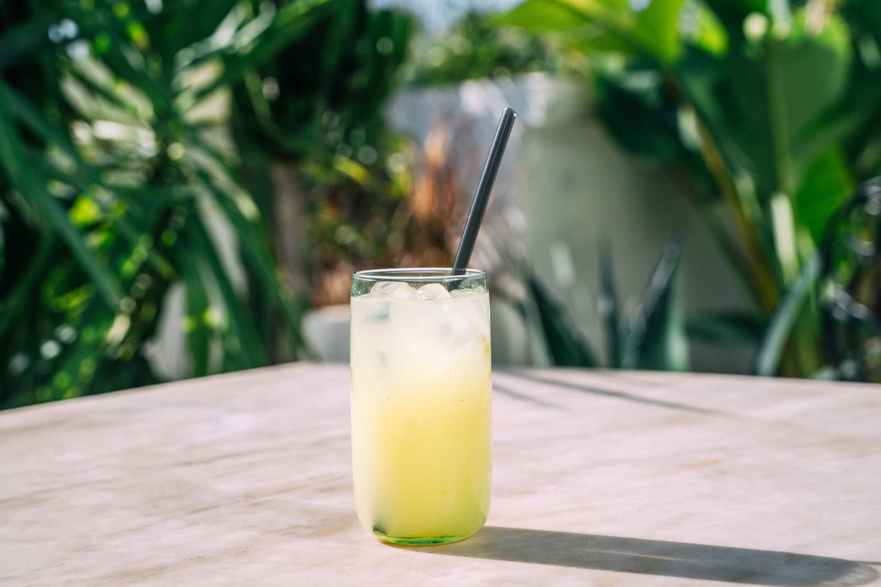 A yellow beverage in a glass over ice with a straw on an outdoor surface.