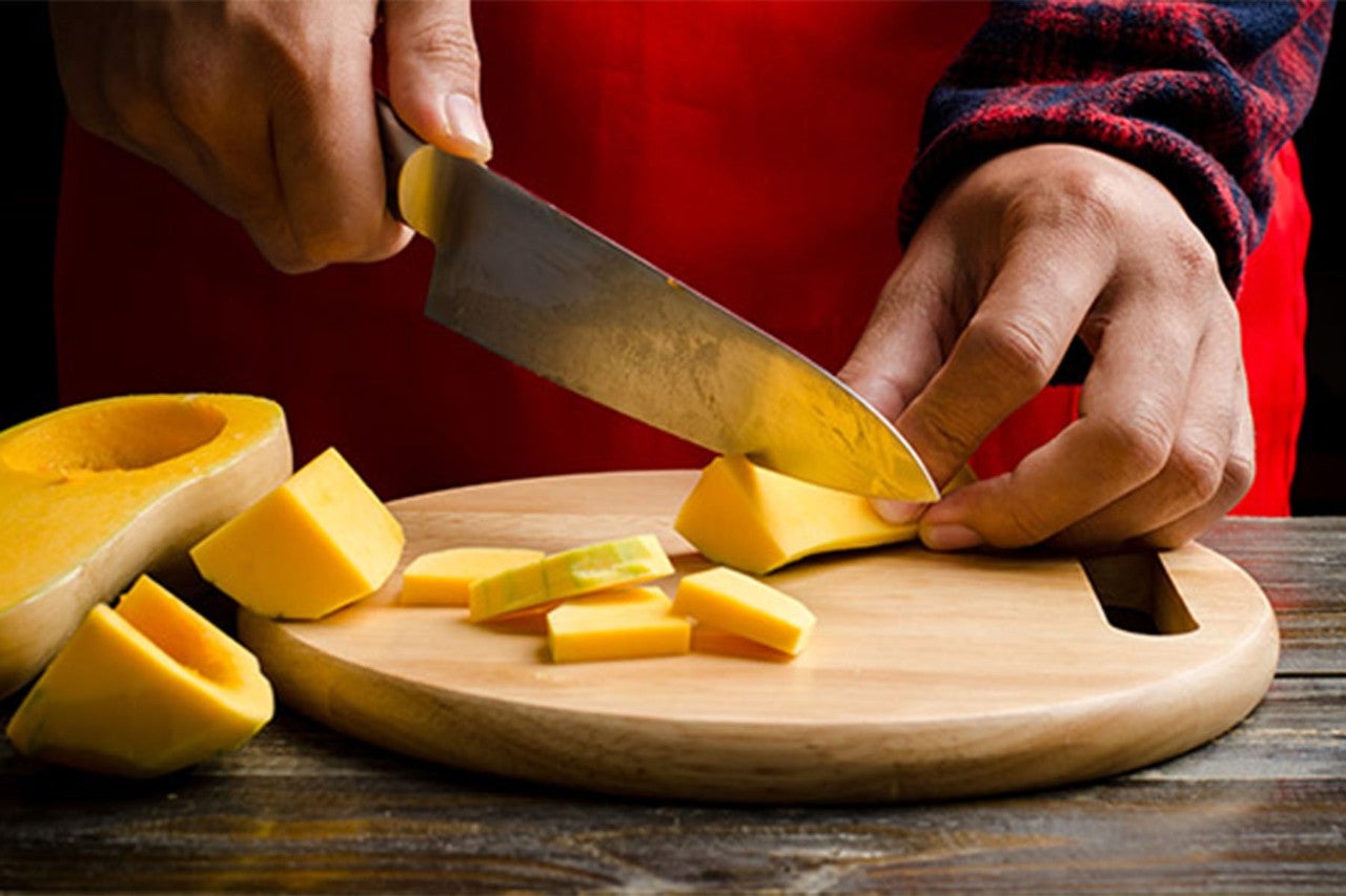 A person in a red apron slicing yellow squash on a wooden cutting board, with pieces of the squash arranged beside it. The background features a rustic wooden table.