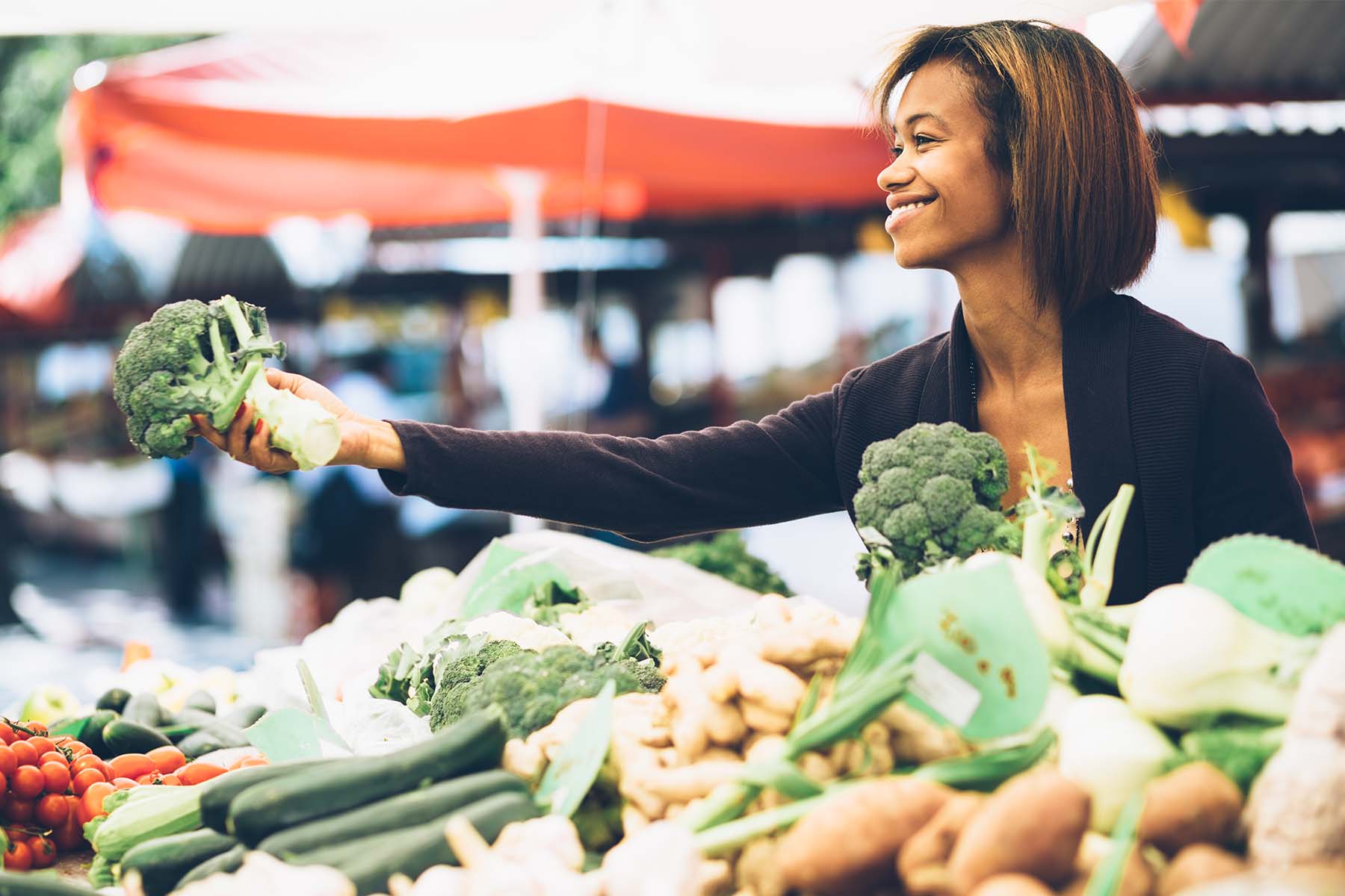 Smiling woman selecting fresh broccoli at an outdoor farmers market, surrounded by a variety of vegetables including zucchini, tomatoes, and ginger.
