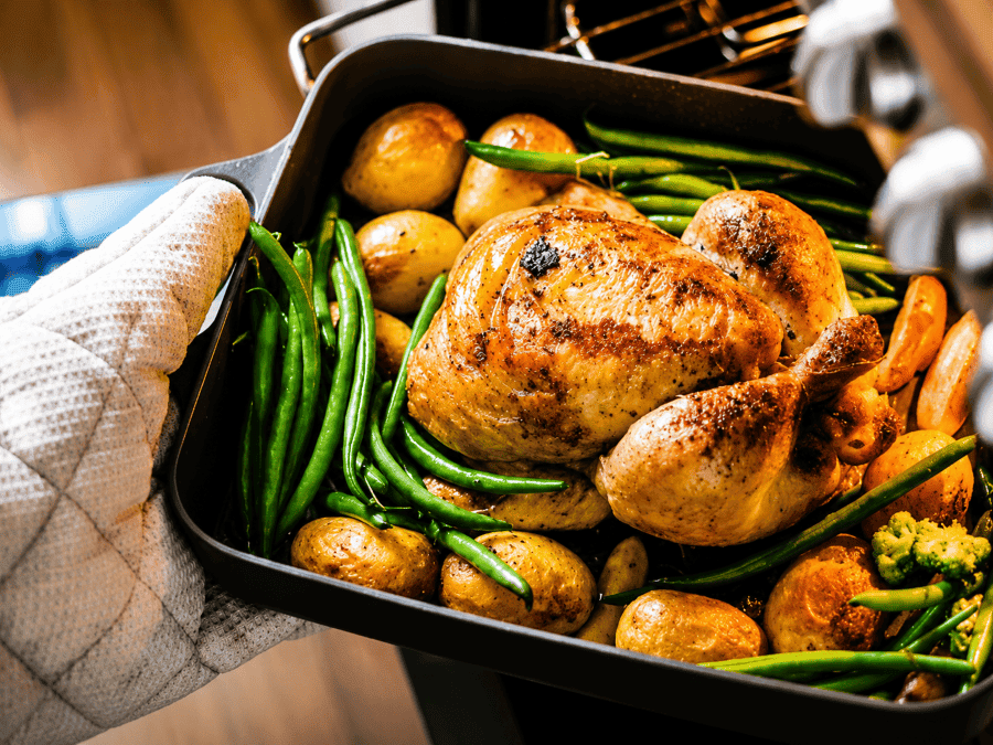 Hand with potholder on it putting a whole chicken in a roasting pan in the oven. The chicken has green beans and potatoes surrounding it..