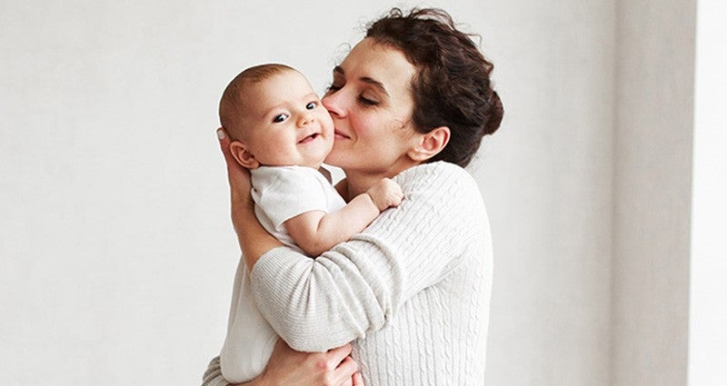 A woman is lovingly holding a baby and giving them a kiss on the cheek, both smiling. The setting is warm and intimate, highlighting their affectionate bond.