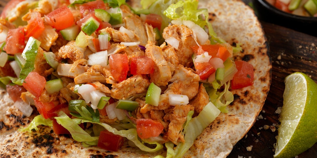 A close-up of a taco filled with shredded chicken, lettuce, diced tomatoes, and chopped green peppers, served on a toasted tortilla, with a lime wedge on the side.