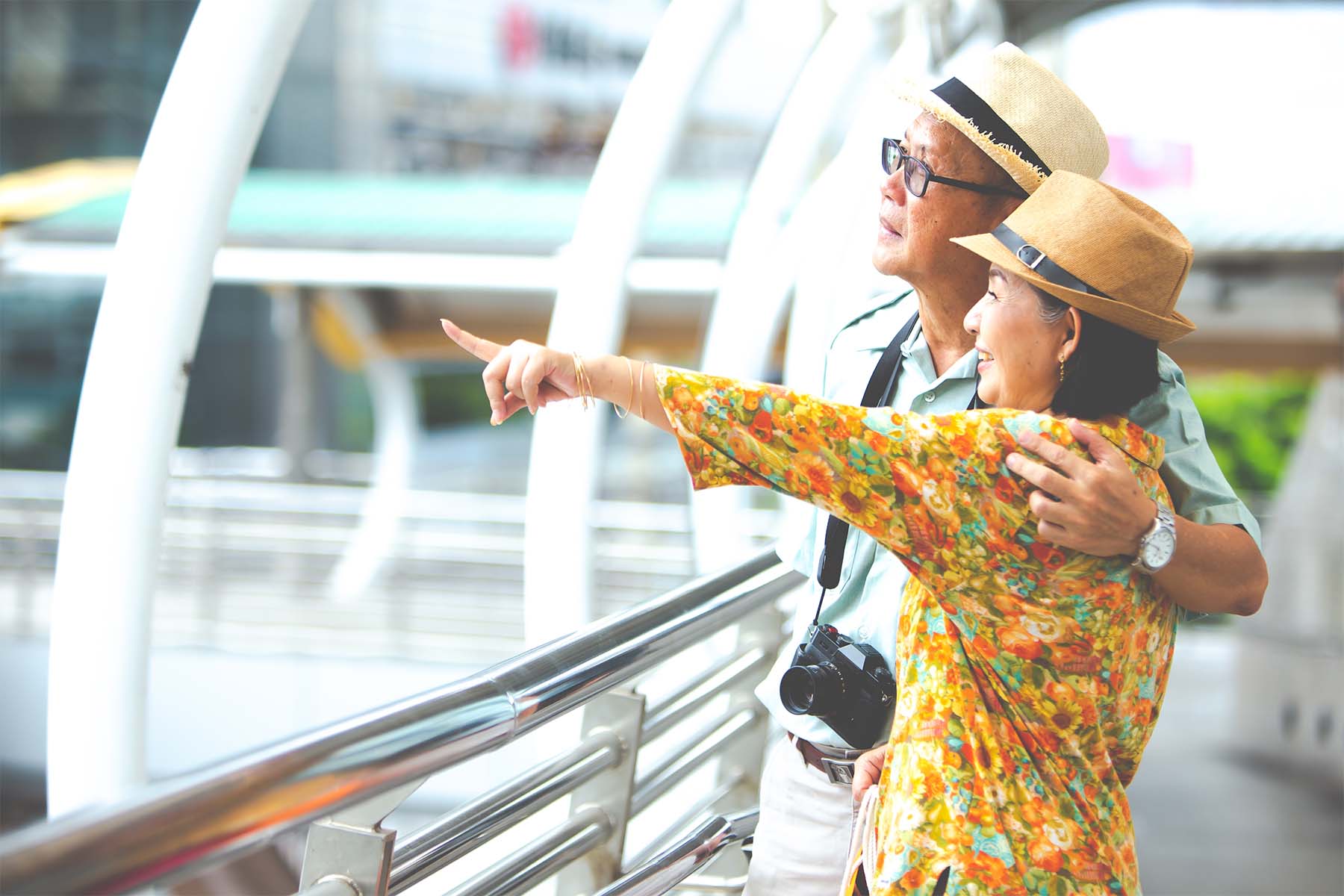 Smiling older couple wearing sun hats, standing by a railing in an urban setting, with the woman pointing at something in the distance and the man holding a camera