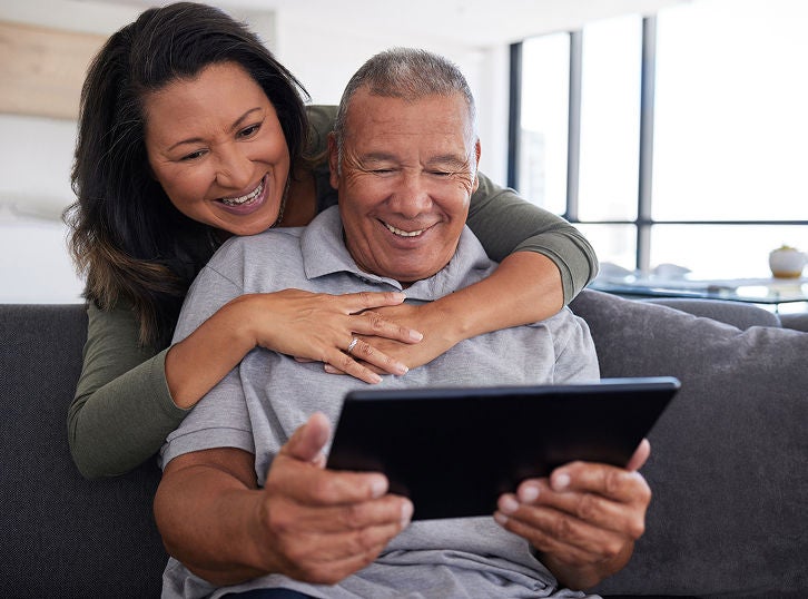 Middle-aged couple sitting on a couch, smiling while looking at a tablet together at home.